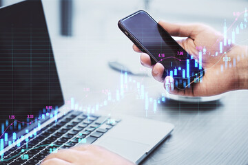 Close up of male hands using laptop and smartphone with glowing upward candlestick forex chart on blurry grid background. Trade, finance and money concept. Double exposure.