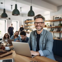 Seated at a table in a well-lit home office, a man exudes happiness and success as he works on his laptop. His smile is evident as he looks directly at the camera.