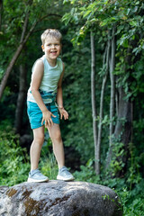 Naklejka na ściany i meble Portrait of little laughing boy stands on big stone in green summer forest.
