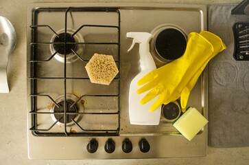 Cleaning a dirty gas surface.  On the stove there is a detergent and cleaning agent, yellow rubber gloves, a rag and a sponge for cleaning the room.  View from above.