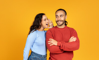 Young woman sharing secret with her husband, yellow background