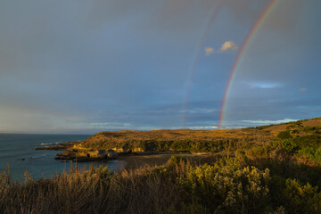 The coastal beach is adorned with the warm hues of a breathtaking sunset, while a vivid rainbow graces the sky