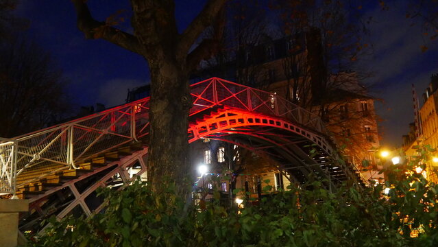 Pont de structure métallique, avec des boulons, marche d'escalier, couple, projection de lumière urbaine rouge ou jaune, en pleine soirée, quartier chic et ancien de Paris, ciel bleu nuageux, traversé
