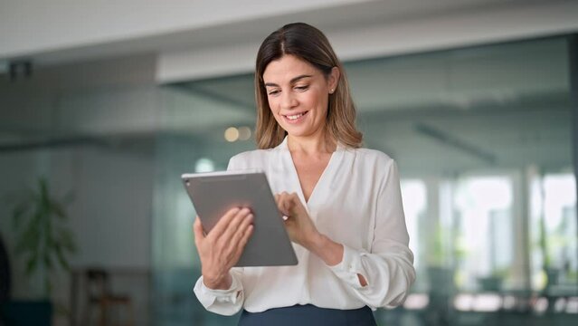 Mid aged busy businesswoman using digital tablet working in office. Middle aged professional business woman company manager standing in office working on corporate tab tech device. - Powered by Adobe