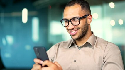 Happy handsome male employee in glasses using mobile phone while sitting in modern glass office at workplace. Close up Young adult businessman typing browsing chatting texting on smartphone Smiling