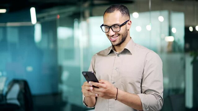 Handsome business man uses a mobile phone, holding in hands. Happy male manager employee in glasses browsing chatting texting using smartphone standing in modern glass office workplace indoor. Smile