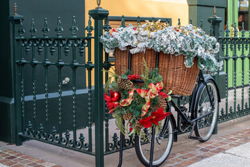 A black two wheel bicycle chained to a dark green wrought iron fence. The butcher's bike has a large wicker basket filled with greenery and Christmas flowers. There's a wreath with red bows on front.