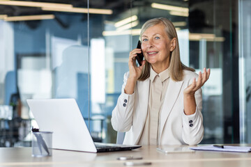 Smiling senior business woman sitting in the office at the desk and smilingly talking on the mobile phone.