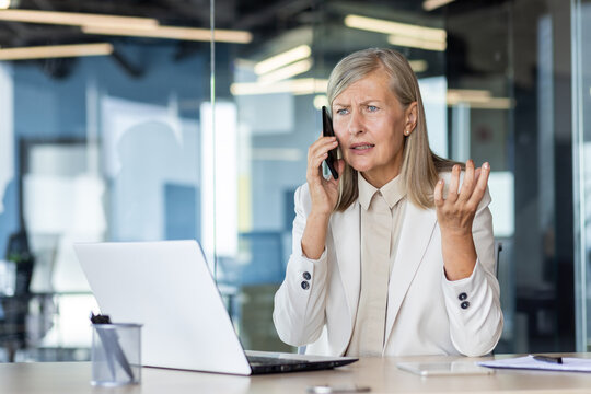 Upset and disappointed senior business woman sitting in the office at the table and talking on the phone.