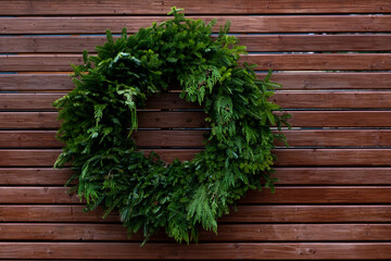 Thick lush green branches in a circular shape with small red berries on a brown fence. The Christmas wreath has natural green fir and cedar boughs. The wall is made of narrow wooden brown boards