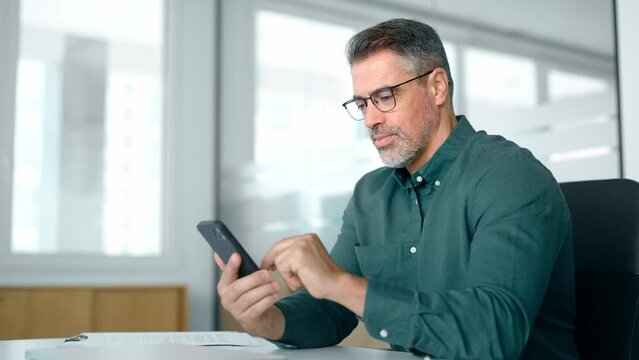 Busy smiling business man executive of mid age looking at smartphone digital technology, older mature businessman entrepreneur investor using cell phone working on mobile cellphone in office.