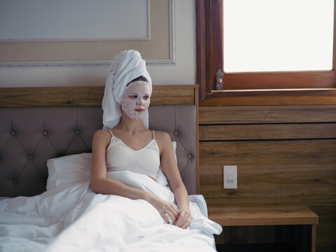 Young woman lying on bed with moisturizing beauty face mask and white towel on her head after shower, lifestyle home skincare, smile.