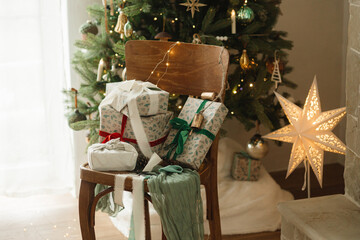Stylish wrapped christmas gifts with ribbon on wooden old chair on background of decorated christmas tree with vintage baubles and festive lights. Merry Christmas and Happy Holidays!