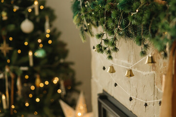Stylish fireplace mantel decorated with christmas branches, bells garland, ornaments on background...