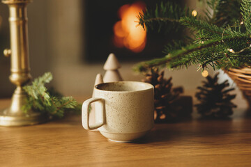 Stylish cup of warm tea, fir branches, wooden trees and star, pine cones on table against burning fireplace. Modern christmas rustic eco friendly decor for winter holidays. Hygge