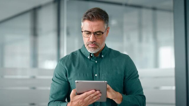 Smiling busy middle aged business man investor using tablet computer, mature older businessman executive manager looking at tab satisfied with finance trading market working standing in office.