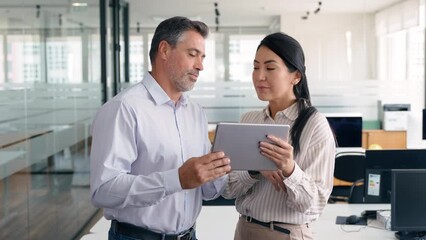 Two happy professional business people team Asian woman and Latin man workers working using digital tablet tech discussing financial market data standing at corporate office meeting. - Powered by Adobe