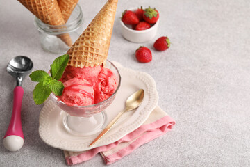 Delicious scoops of strawberry ice cream with mint and wafer cone in glass dessert bowl served on grey table. Space for text