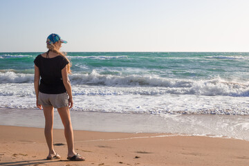 A woman with long blond hair walks along the sandy seashore on a windy day. Travel and tourism.