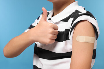 Boy with sticking plaster on arm after vaccination showing thumbs up against light blue background,...