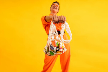 woman wear modern orange overalls with eco string bag and colored gingerbread cookies on bright yellow color background. homemade colored dessert