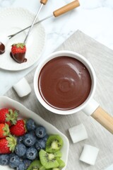 Fondue pot with melted chocolate, sweet marshmallows, fresh kiwi, different berries and forks on white marble table, flat lay