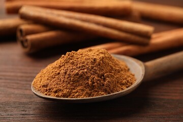 Spoon with cinnamon powder and sticks on wooden table, closeup