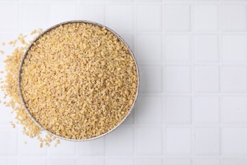 Raw bulgur in bowl on white tiled table, top view. Space for text