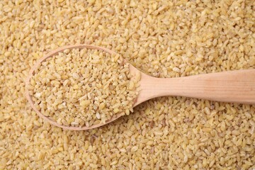 Wooden spoon and raw bulgur as background, top view
