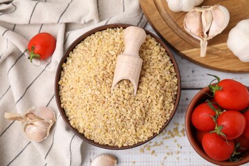 Bowl of raw bulgur with scoop, tomatoes and garlic on white wooden table, flat lay