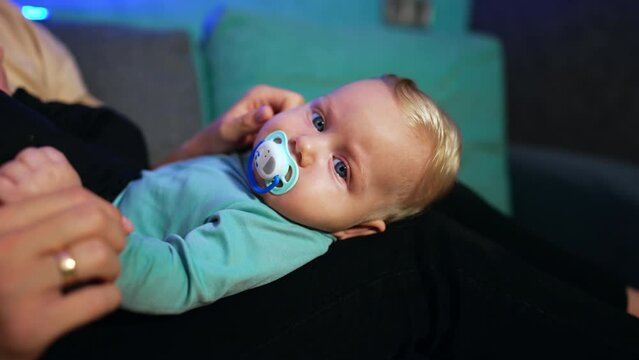 Lovely child with fair hair lies on mom's laps. Mother takes a pacifier out of kid's mouth and waves it in front of child's face. Close up.
