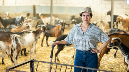 Glad smiling professional young Latin woman farmer standing with pitchfork during work on goat...