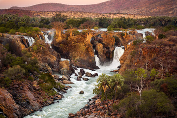 Epupa Falls, Kunene Region, Namibia, in warm, golden light. Epupa Falls is a series of large waterfalls formed by the Kunene River on the border of Angola and Namibia. - obrazy, fototapety, plakaty