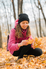 Portrait of beautiful happy young woman in pink jacket holding bouquet of yellow leaves at autumn park. Pretty Caucasian lady smiling and looking at camera during her walk outdoors. Generation Z and