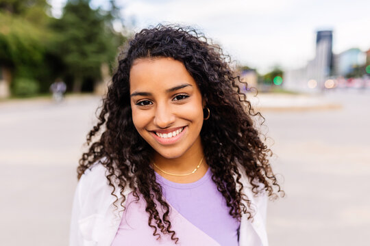 Happy young latin american girl smiling at camera standing at city street. Outside portrait of joyful beautiful woman over urban background. - Powered by Adobe
