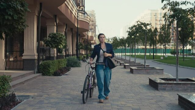 A woman finishes her coffee before work while walking with a bicycle in the cityscape. a businesswoman rushes to the office of the business center on a bicycle. High quality 4k footage