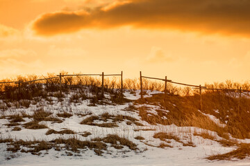 A dramatic orange evening sky with dark clouds over a grassy hill covered with grass, hay and white fresh snow in a meadow. There's an old wooden farm fence on the hillside with a small opening.  - Powered by Adobe