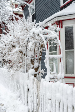 The exterior entrance to a blue colored Victorian style building with red and white trim on the bay window. There's a white wooden picket fence and arbor or archway covered in fresh white winter snow.