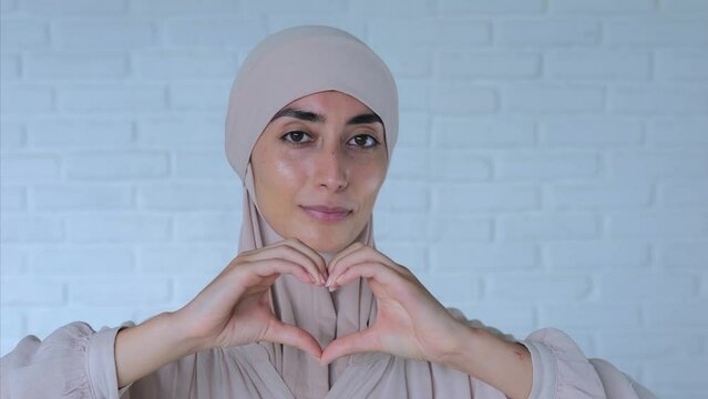 Beautiful Muslim woman shows heart with hands and smiles at camera. Cinematic portrait of lady in hijab shows us heart with hands and sharing love. Concept of having inner love in heart to world