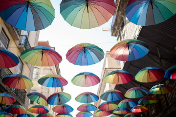 Fototapeta na wymiar Colorful floating umbrellas hang above the street. Street decorated with colored parasols. Rainbow umbrellas on blue sky background. Many rainbow parasols, street decoration for festivals. Pride month