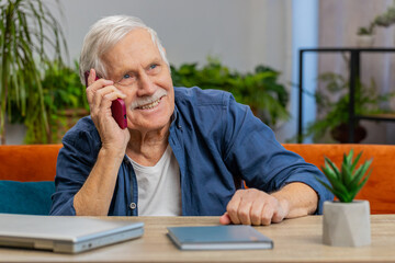 Phone call, good news, gossip. Happy smiling surprised old senior man in pleasant conversation on smartphone, enjoying talking with friend at home apartment. Elderly grandfather sits on couch at table