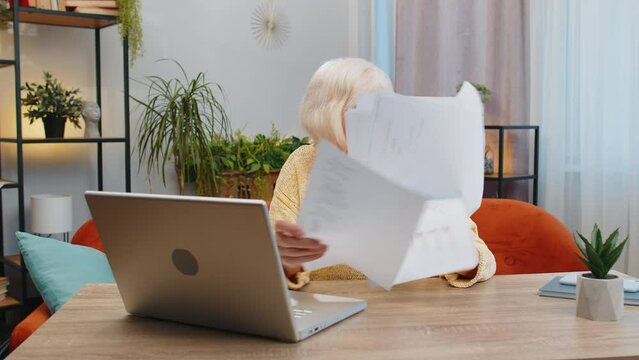 Stressed senior woman at home room looking at unpaid bank debt bills, doing paperwork, planning budget calculate finances mortgage payments. Elderly mature grandmother throws paper bills sits at table