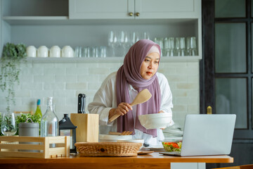 muslim mother looking at recipe from laptop and cooking. having fun woman with hijab preparing dinner, Islamic woman Enjoying Doing Homemade, Teaching cooking online