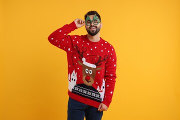 Happy young man in Christmas sweater and funny glasses on orange background