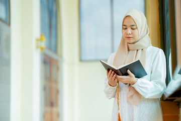 The image of an Asian Muslim woman in the Islamic religion in hijab in cream color. reading the Quran and having a happy Staying in a beautiful mosque, Arabic word translation: The Holy Al Quran.