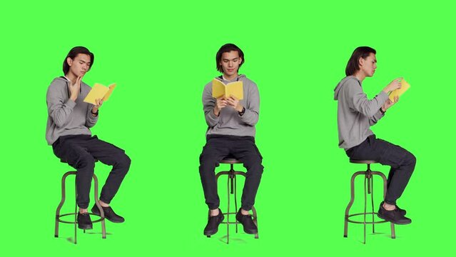 Trendy person reading novel book on chair against full body greenscreen template, enjoying old school lecture hobby. Young asian adult holding literature or fiction book to read in studio.