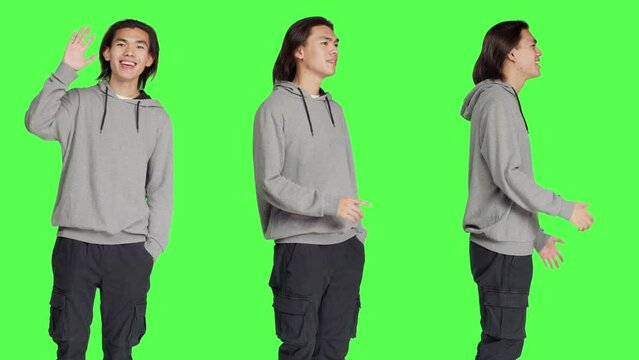 Asian guy waving hello at someone, greeting people with a smile and confidence against greenscreen template. Young adult saying hi with a wave, smiling and feeling cheerful in studio.