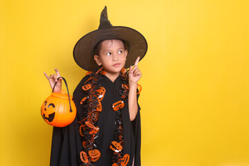 Young Asian little girl in witch costume to celebrating Halloween pointing up to empty space for text or ads while holding orange pumpkin bucket 