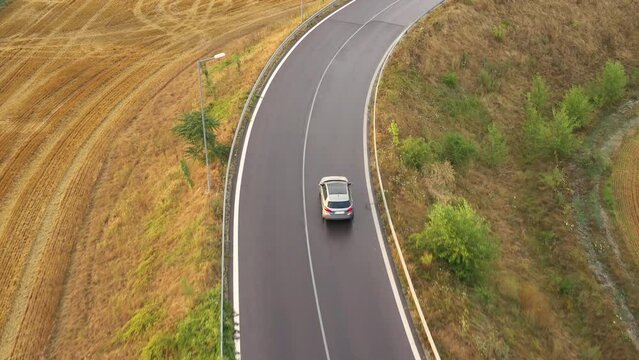Tracking a silver car on the asphalt road - aerial done shot. A silver car drives through the countryside at sunny day. Yellow field by the road. New asphalt road.