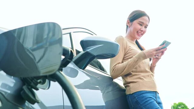 Asian woman driving an electric car checks cables while charging the battery and easily takes time stand and relax while playing with her smartphone while waiting for the battery charge short time.